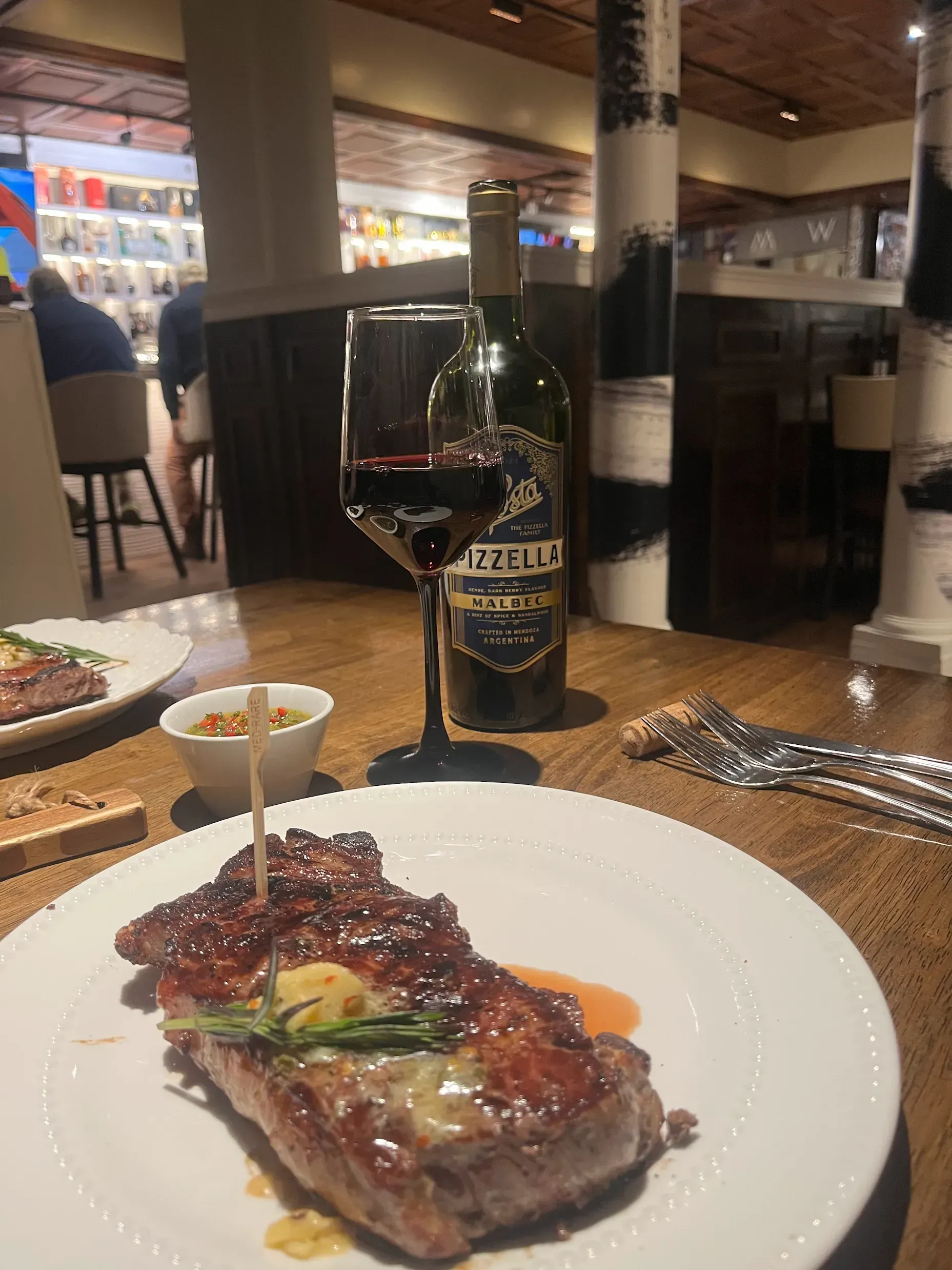 A steak dinner with a glass and bottle of red wine served at a restaurant table.