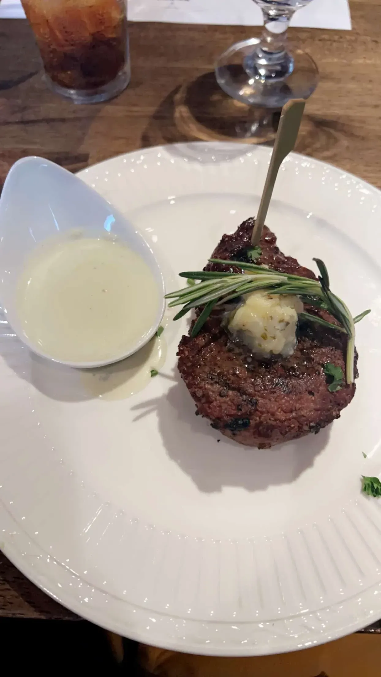 A seared steak topped with a pat of butter and herbs, served with a small side of white sauce on a white plate.
