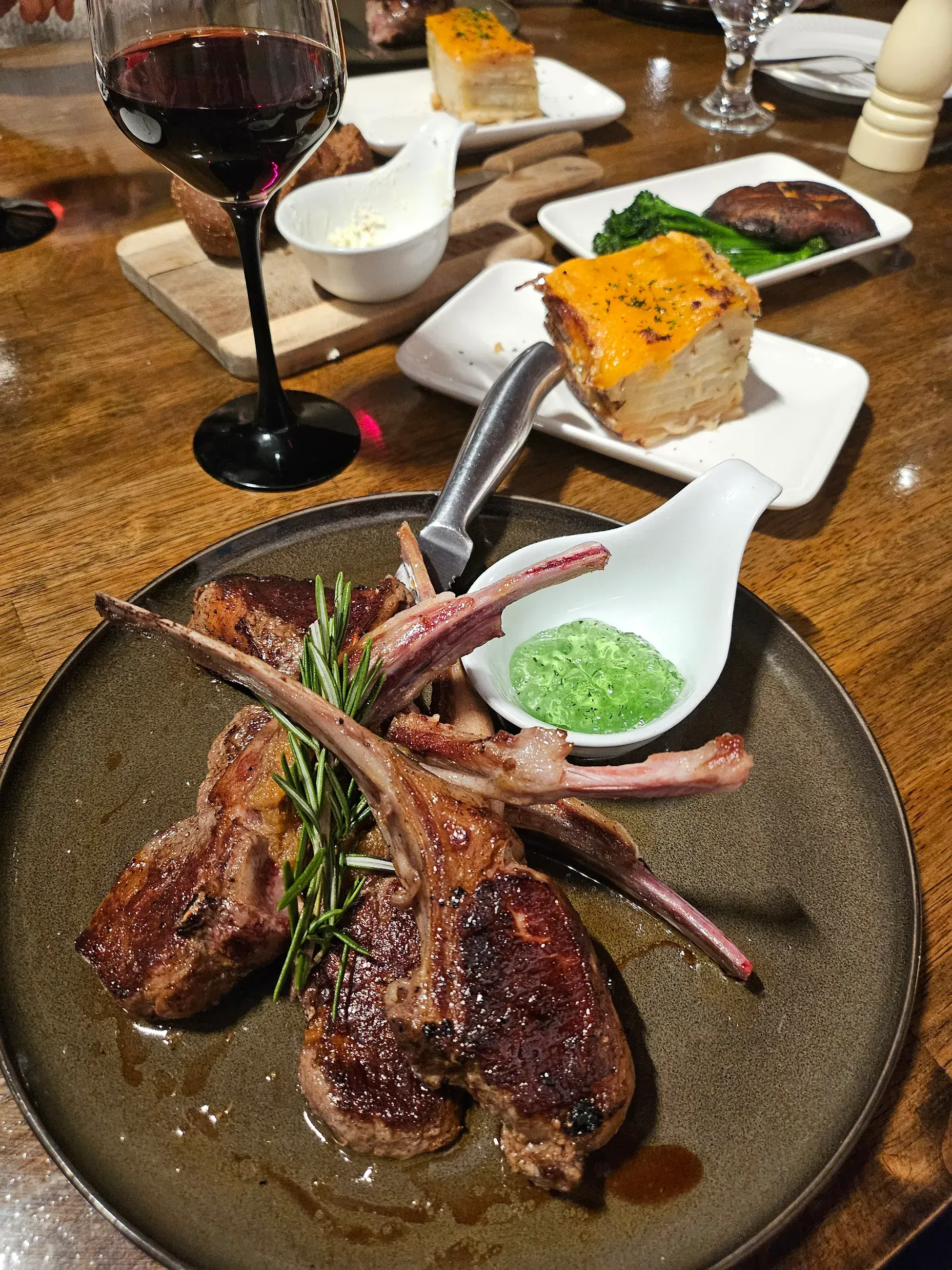 A grilled lamb chop dinner with a side of green herb sauce, a square potato gratin, and a glass of red wine on a table.