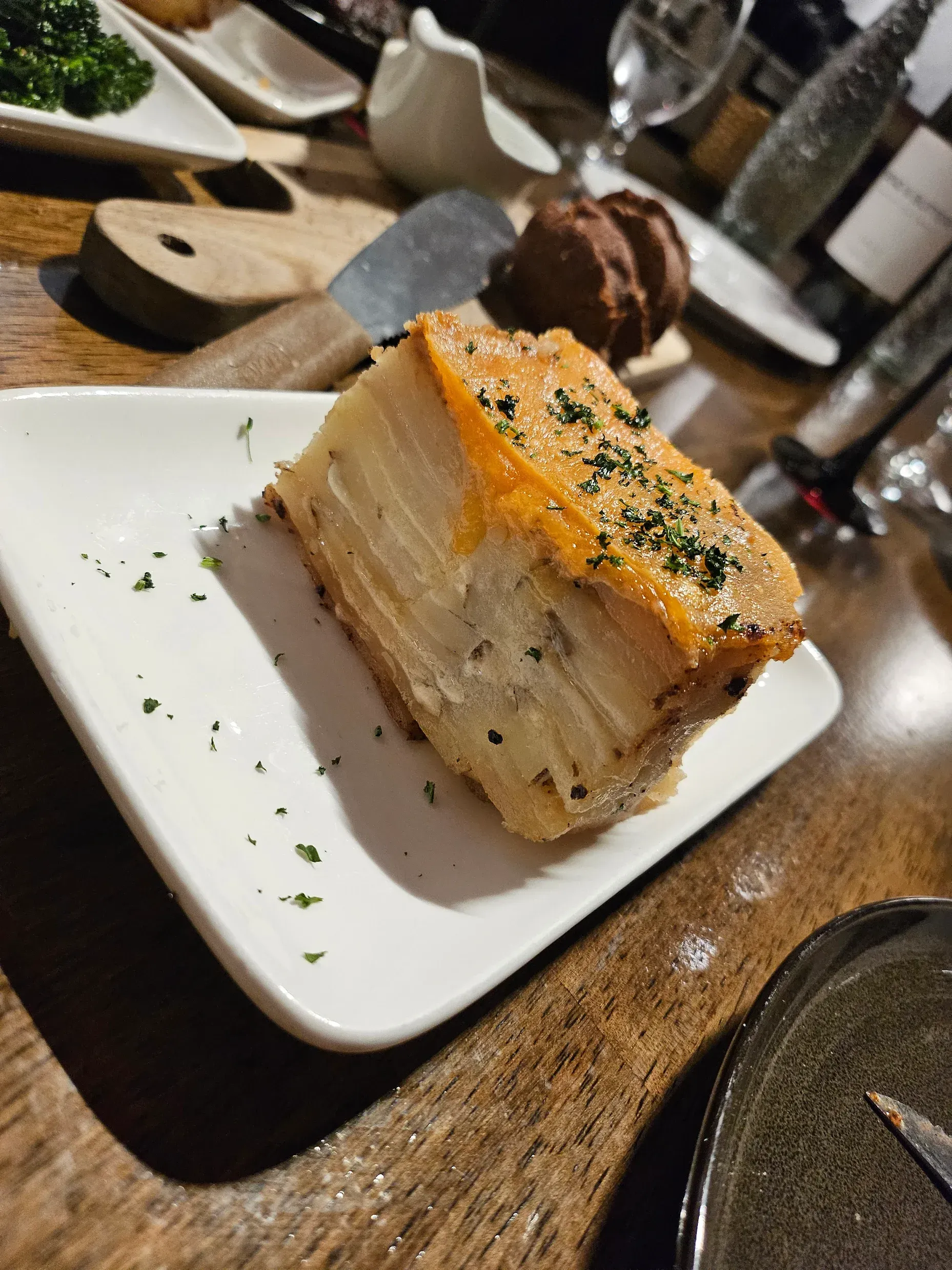 A block of layered potato gratin topped with melted cheese and fresh herbs, served on a white plate on a wooden table.