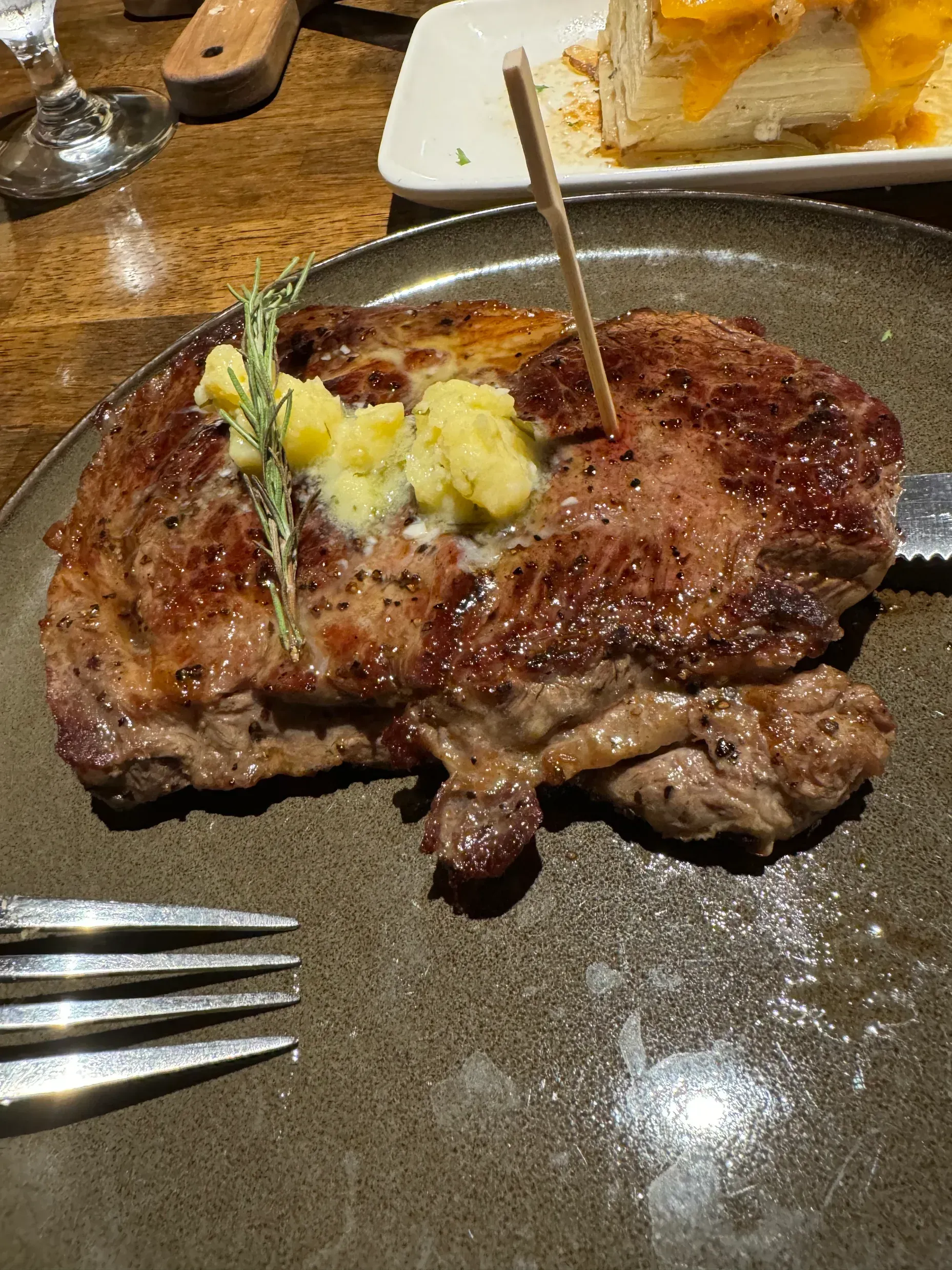 A grilled steak topped with melting butter and a sprig of rosemary on a dark plate, with a fork and side dish nearby.