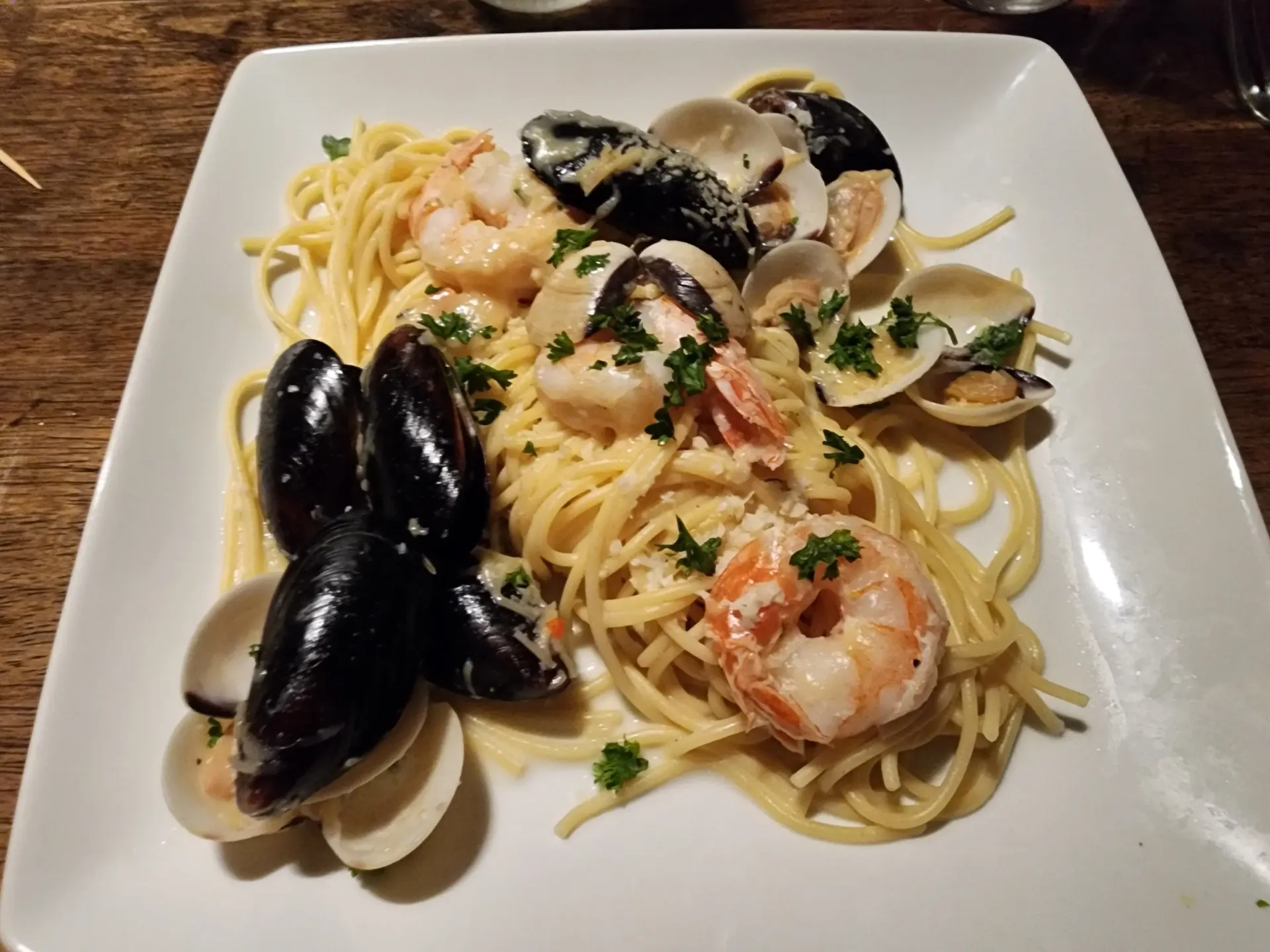 A plate of seafood pasta featuring mussels, clams, and shrimp served over a bed of linguine on a white square plate.