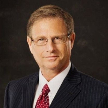 Man in a suit and tie, wearing glasses, smiling slightly, against a dark background.