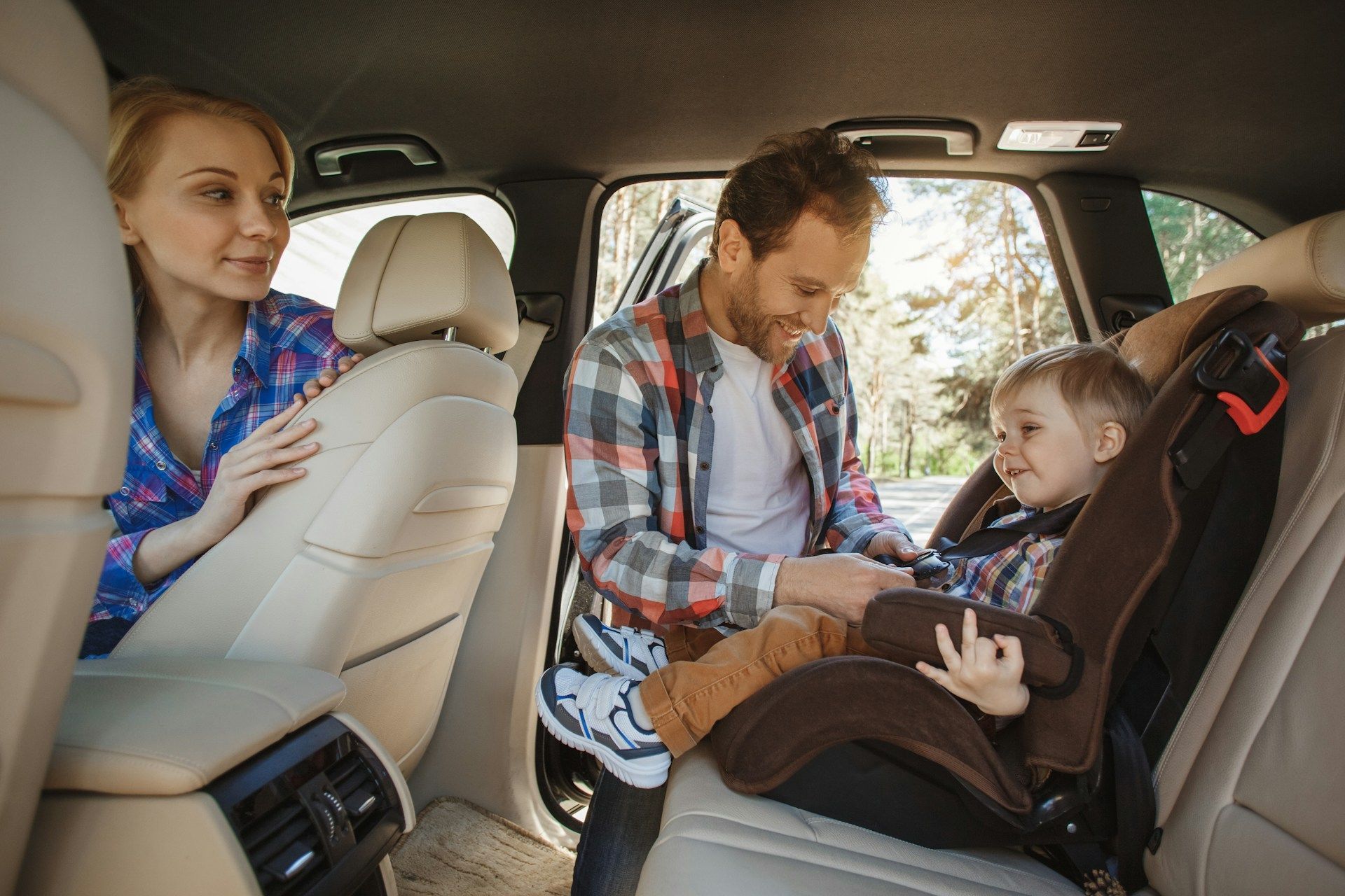 Family in car, father securing toddler in a car seat, mother looking on.