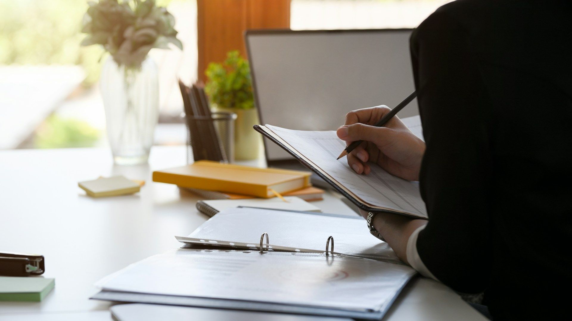 Person in a blazer writing on a clipboard at a desk with a laptop, books, and plants in the background.