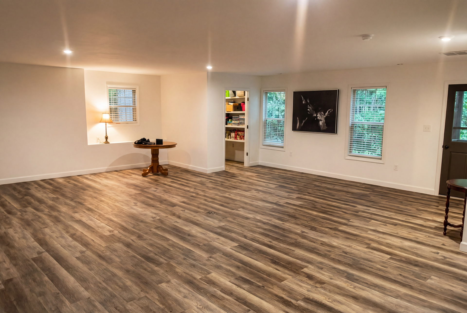 Empty modern living room with wood flooring, recessed lights, windows, and a small table near the wall