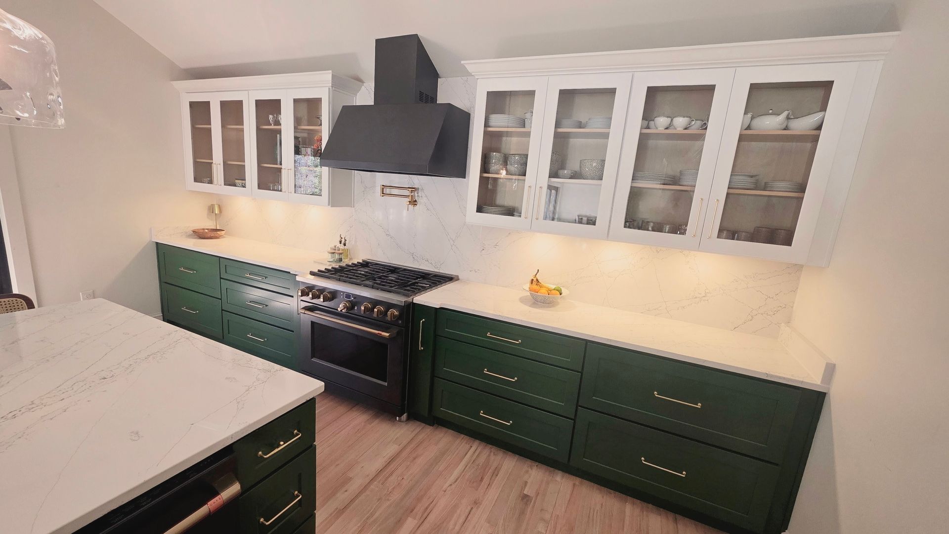 Green and white kitchen with dark range hood and glass-front cabinets above dark green cabinets and white countertops.