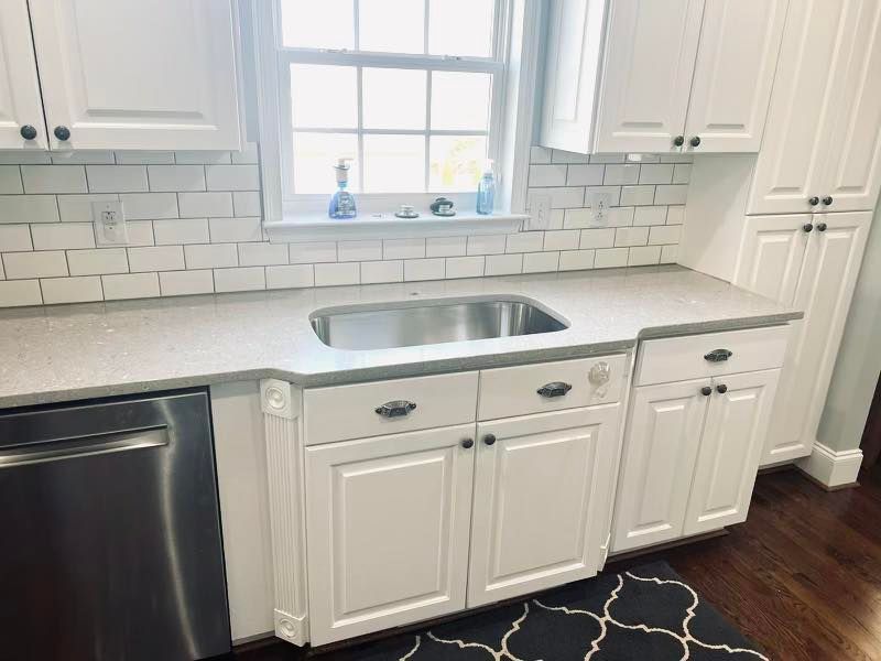 White kitchen with sink under a window, white cabinets, subway tile backsplash, and dark wood floor.