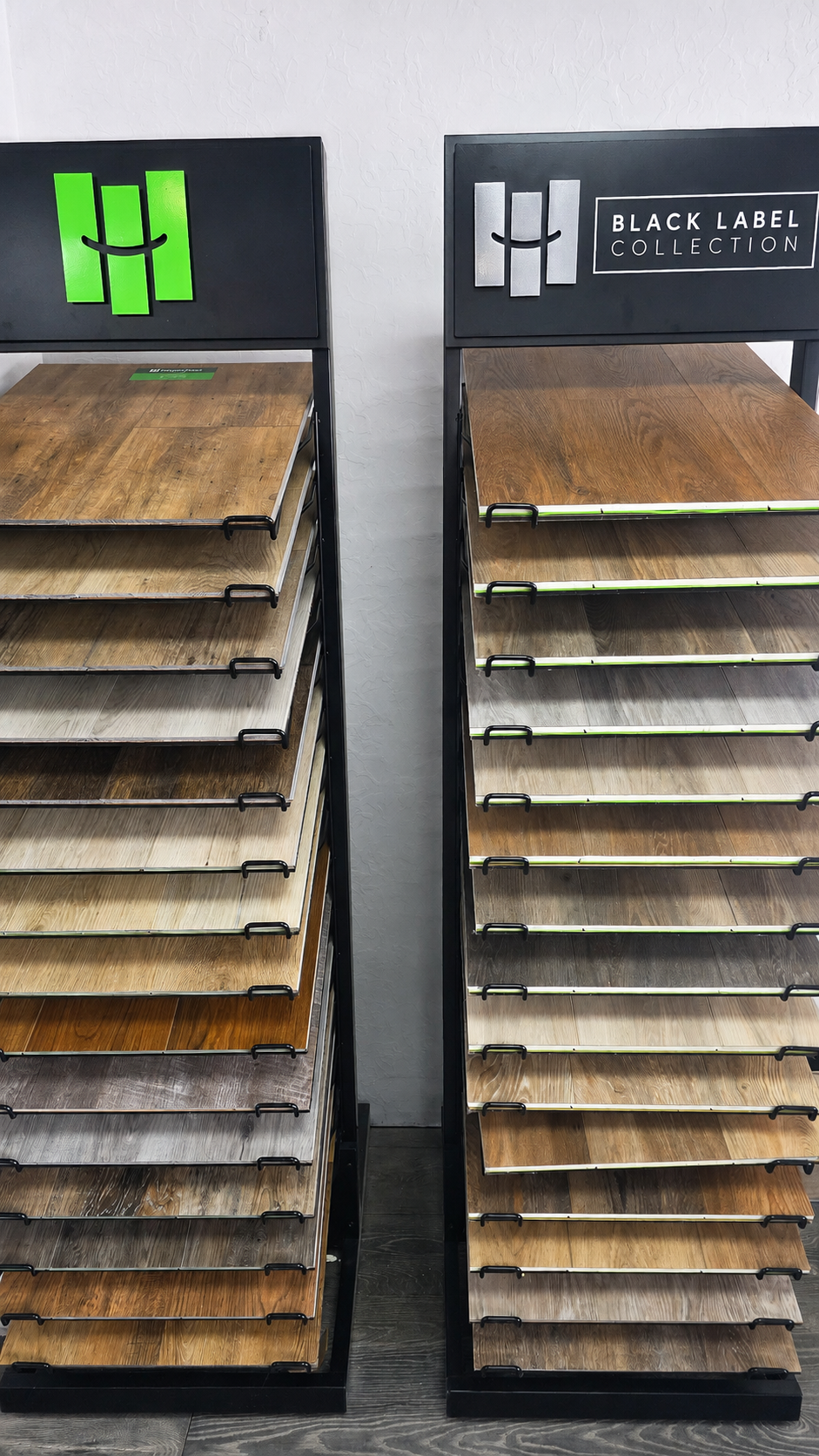 Two sample flooring display racks with stacked wood planks, one with a green logo and one with a black sign.