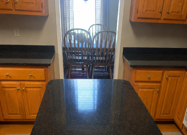 Kitchen with black countertops and wooden cabinets, looking through to a dining area with chairs and bright window.