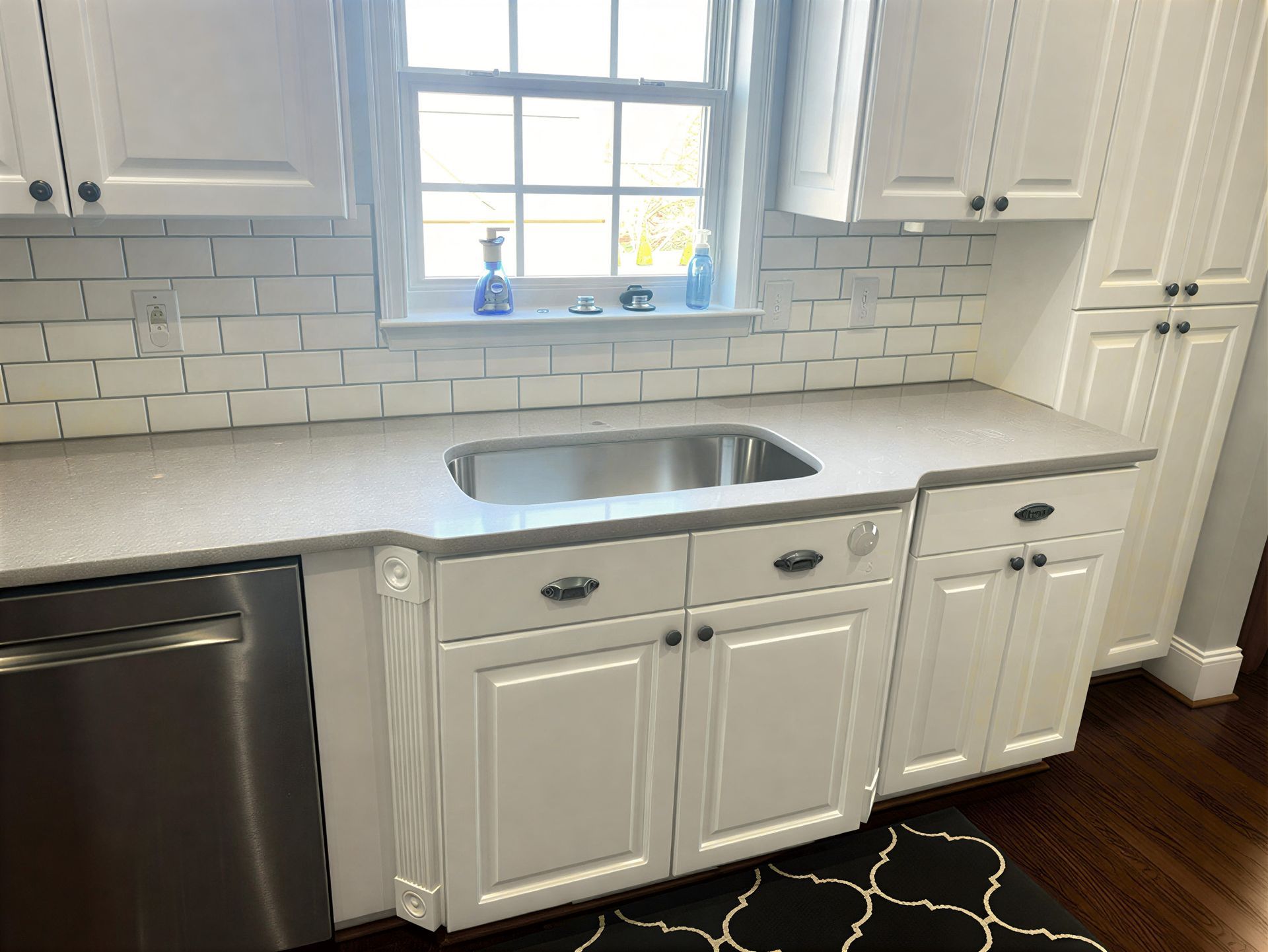 White kitchen cabinets and counters with a stainless steel sink, tile backsplash, and window.