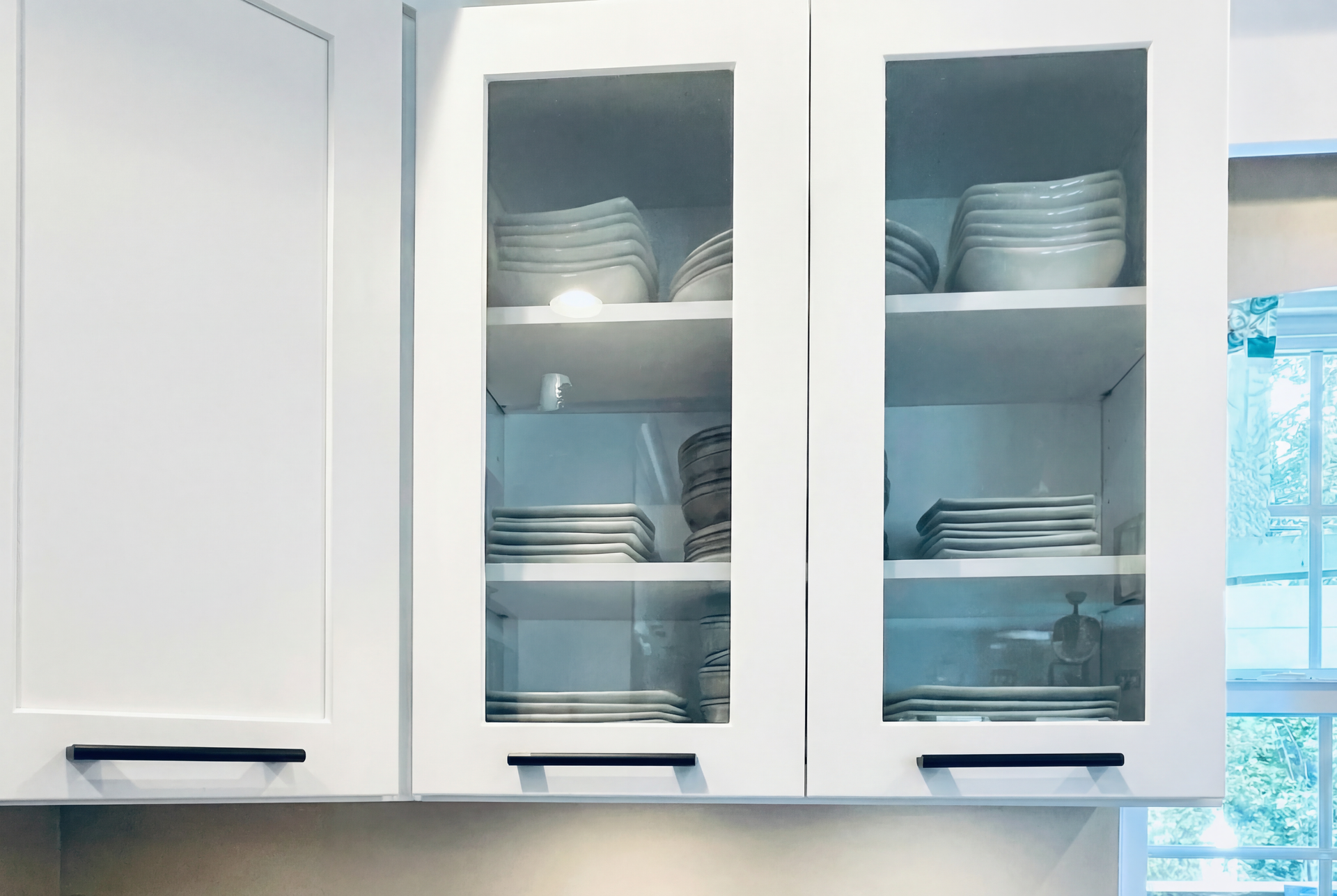 White kitchen cabinets with glass-fronted center doors displaying dishes. Black handles below.