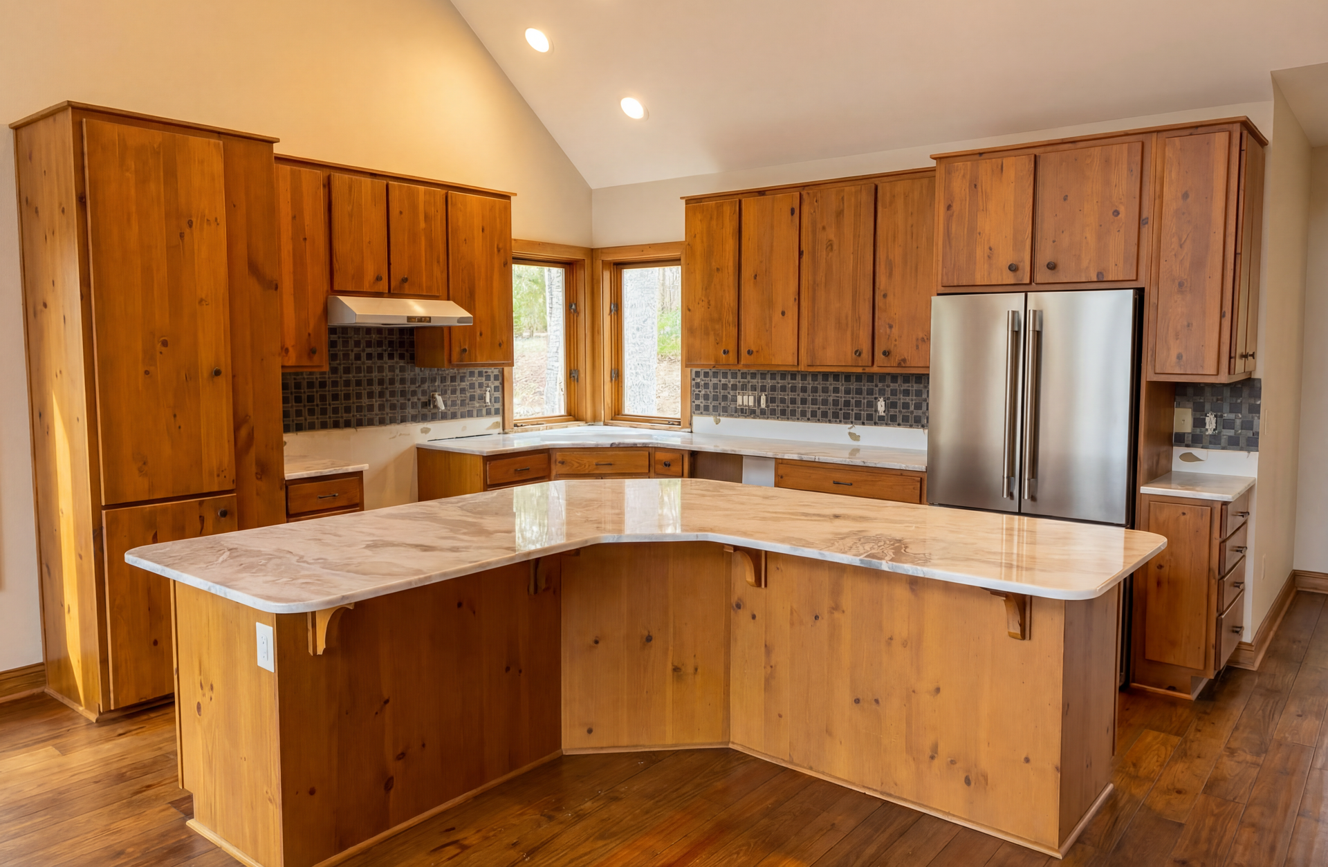 Kitchen with light-brown wooden cabinets, white countertops, and a curved island. Stainless steel refrigerator, and recessed lighting.