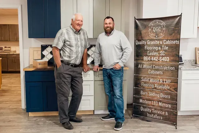 Two men stand in a kitchen beside a wooden sign with white lettering.