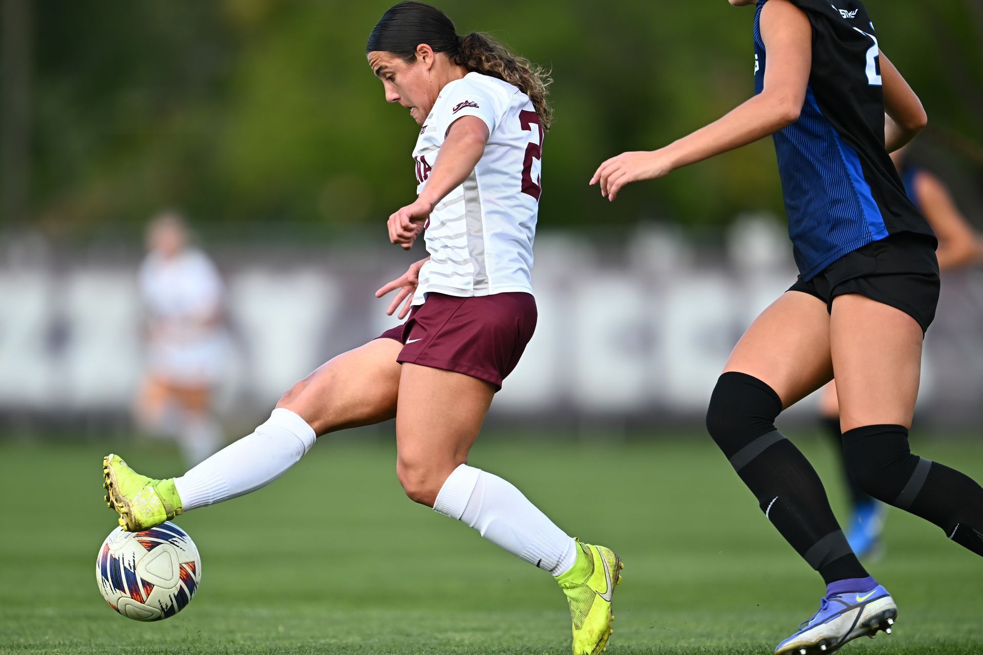 A soccer player in a white jersey and maroon shorts kicks a ball on a green field as a player in a blue jersey defends.