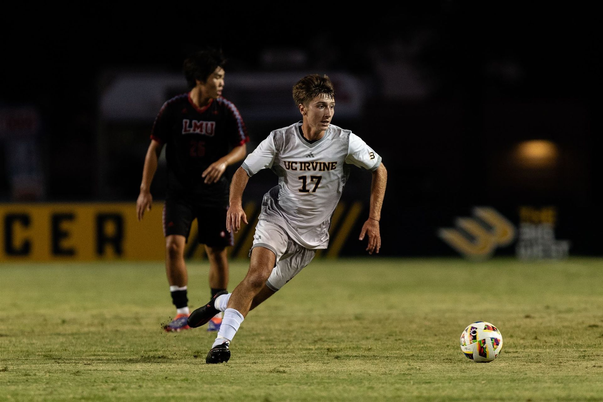 A soccer player in a white uniform with number 17 dribbles a ball on a grass field, with an opponent in dark gear nearby.