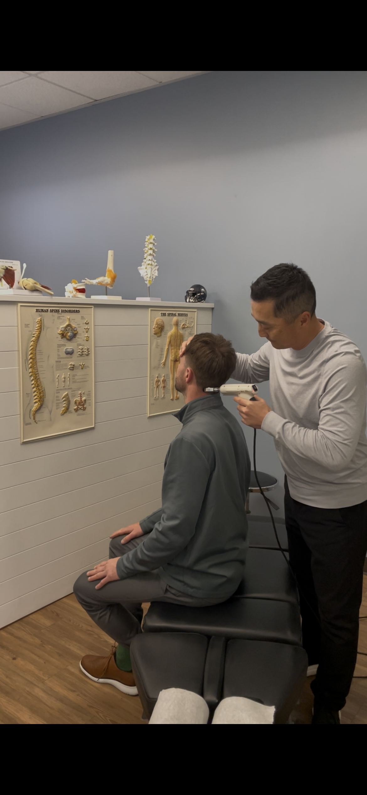 A practitioner uses an adjusting tool on a patient's neck in a clinical office setting with anatomical charts displayed.