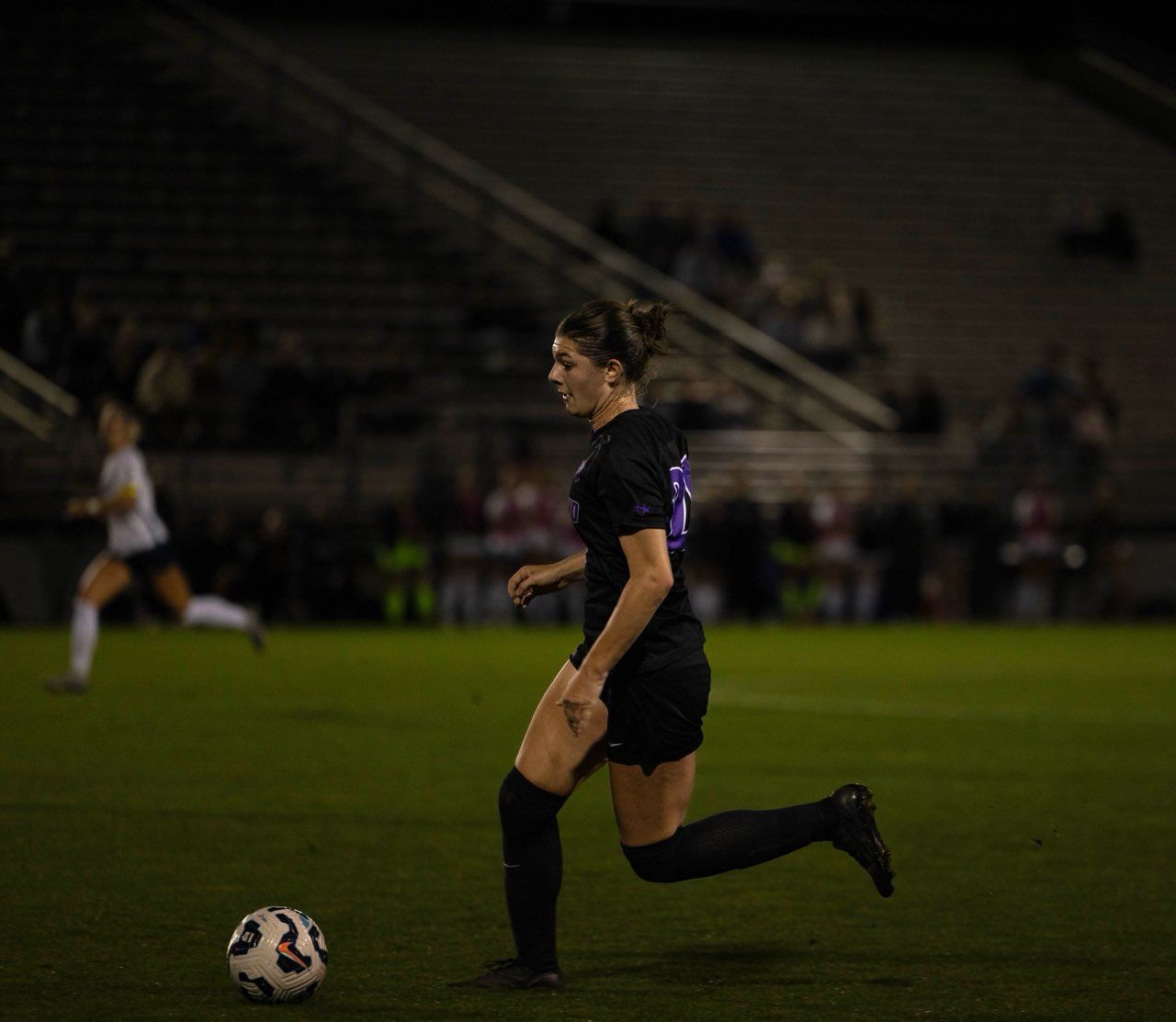 A soccer player in a black uniform dribbles a ball on a lit field at night, with another player blurred in the background.