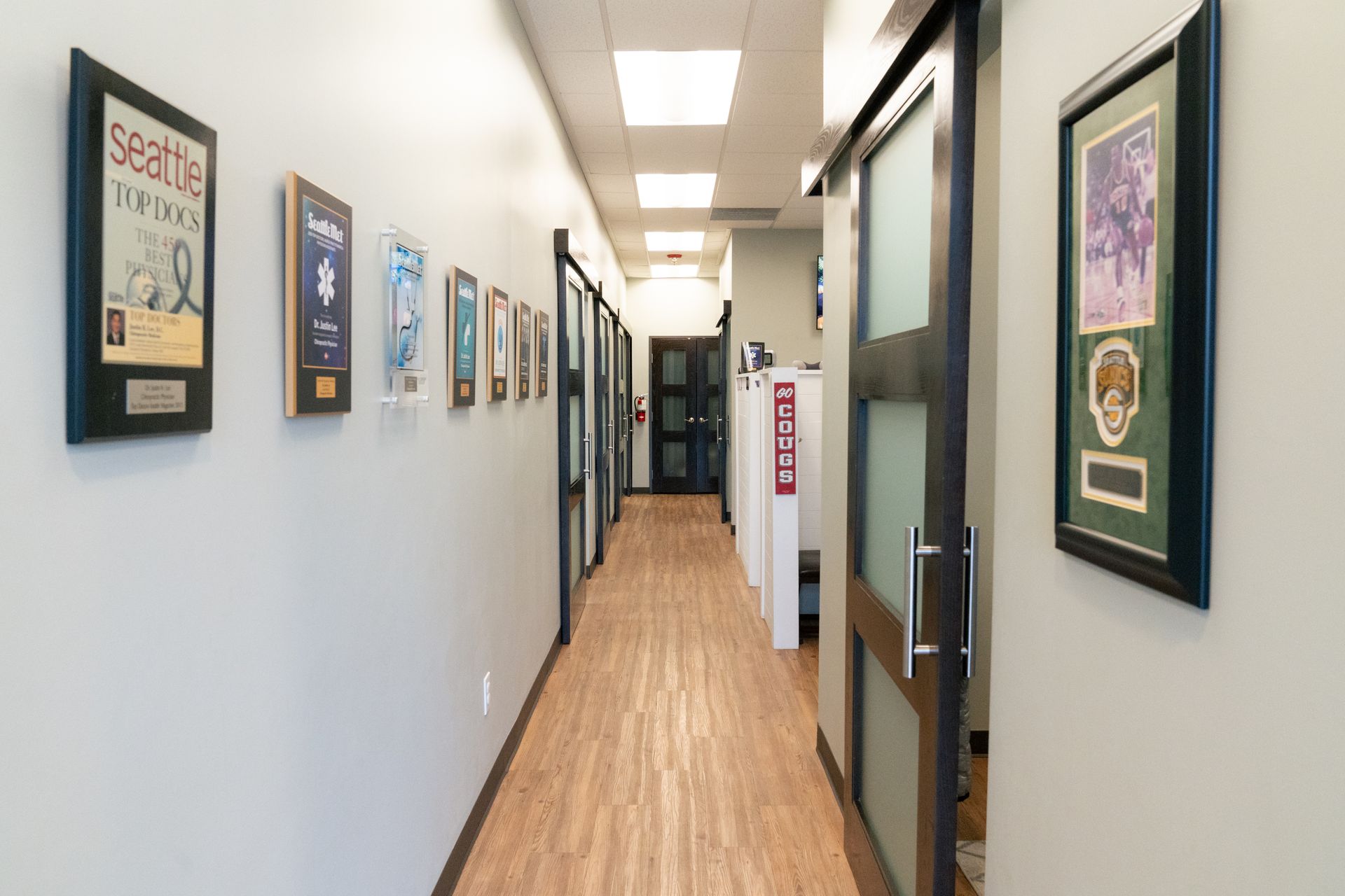 A well-lit office hallway with light wood floors, framed awards on the left wall, and sliding glass doors to workspaces.