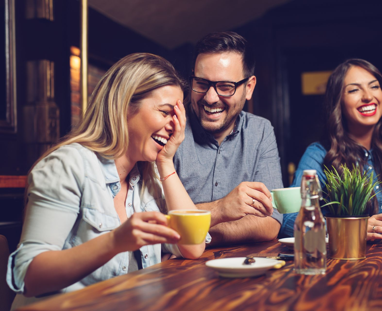 Three people laughing and drinking coffee at a wooden table in a dimly lit cafe.