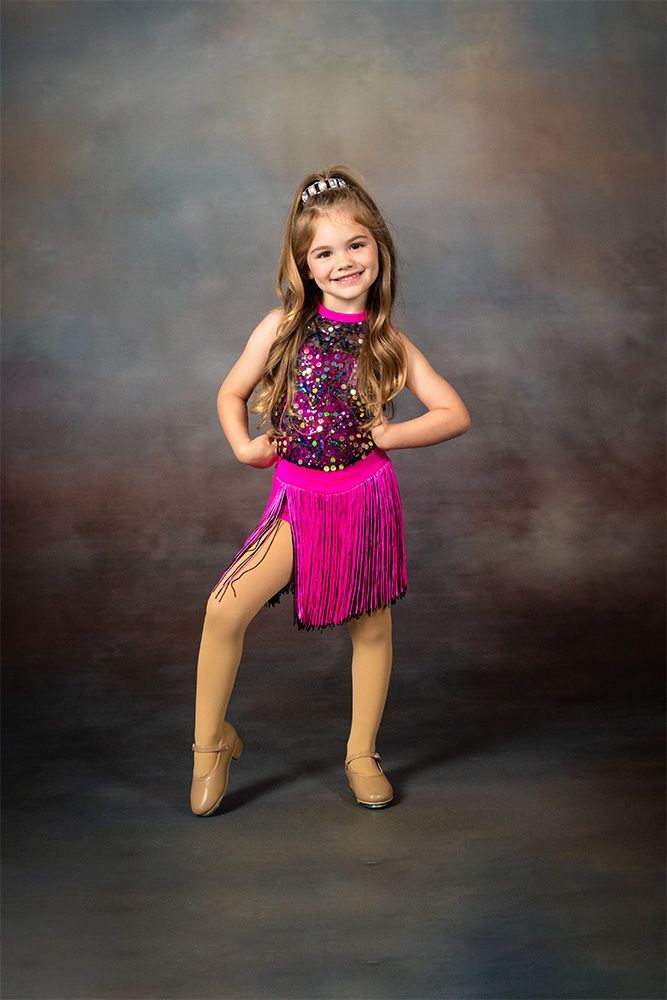 Young dancer in a pink sequined costume posing with one hand on her hip against a studio backdrop