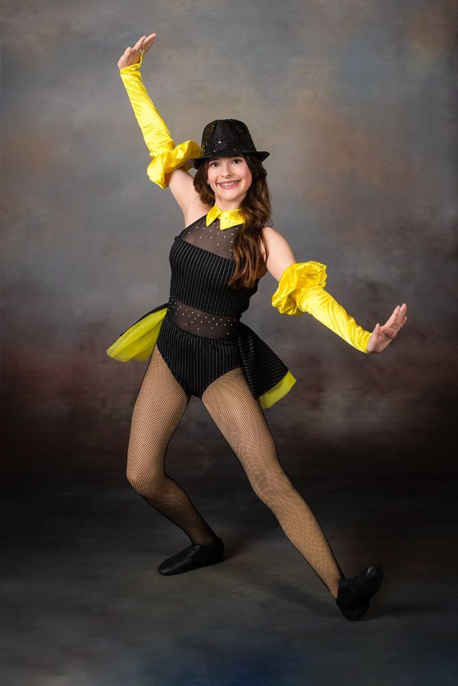Dancer in black costume with yellow accents, posing with arms raised against a studio backdrop