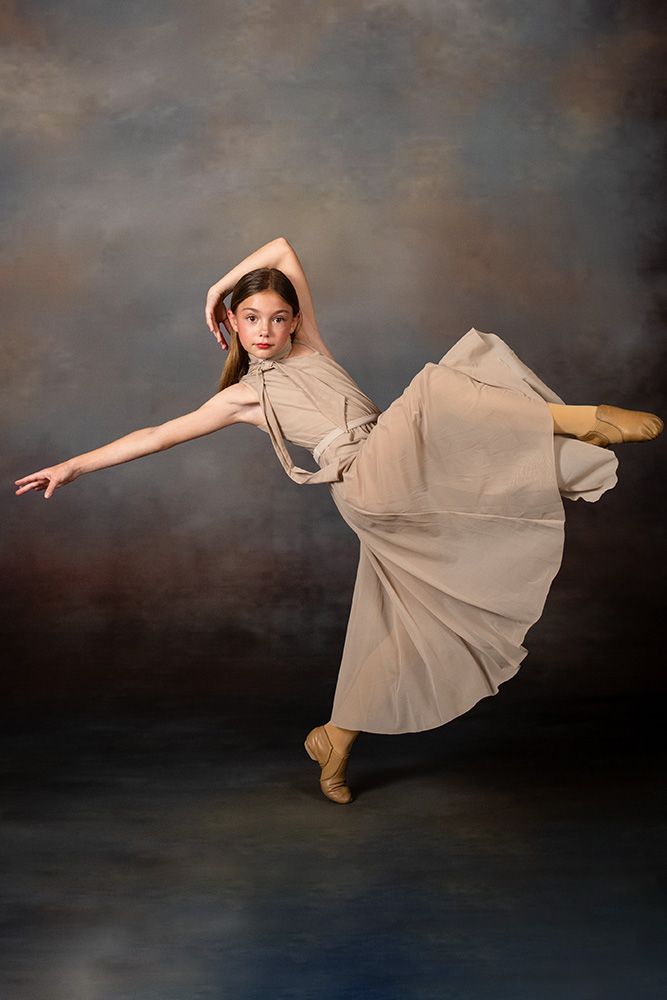 Dancer in a beige dress and boots posing mid-kick against a dark studio backdrop