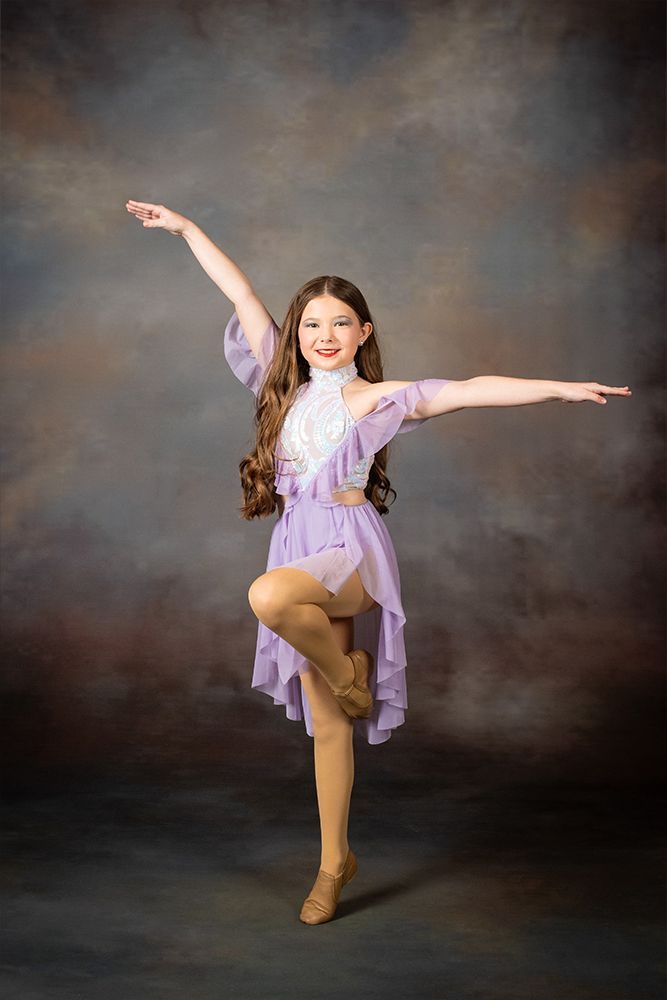 Young dancer in a lavender dress posing on one leg with arms outstretched against a dark studio backdrop