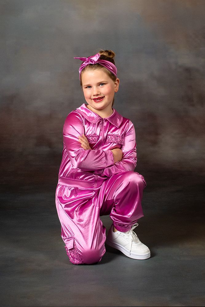 Child in shiny pink outfit kneeling with arms crossed, smiling against a gray studio backdrop.
