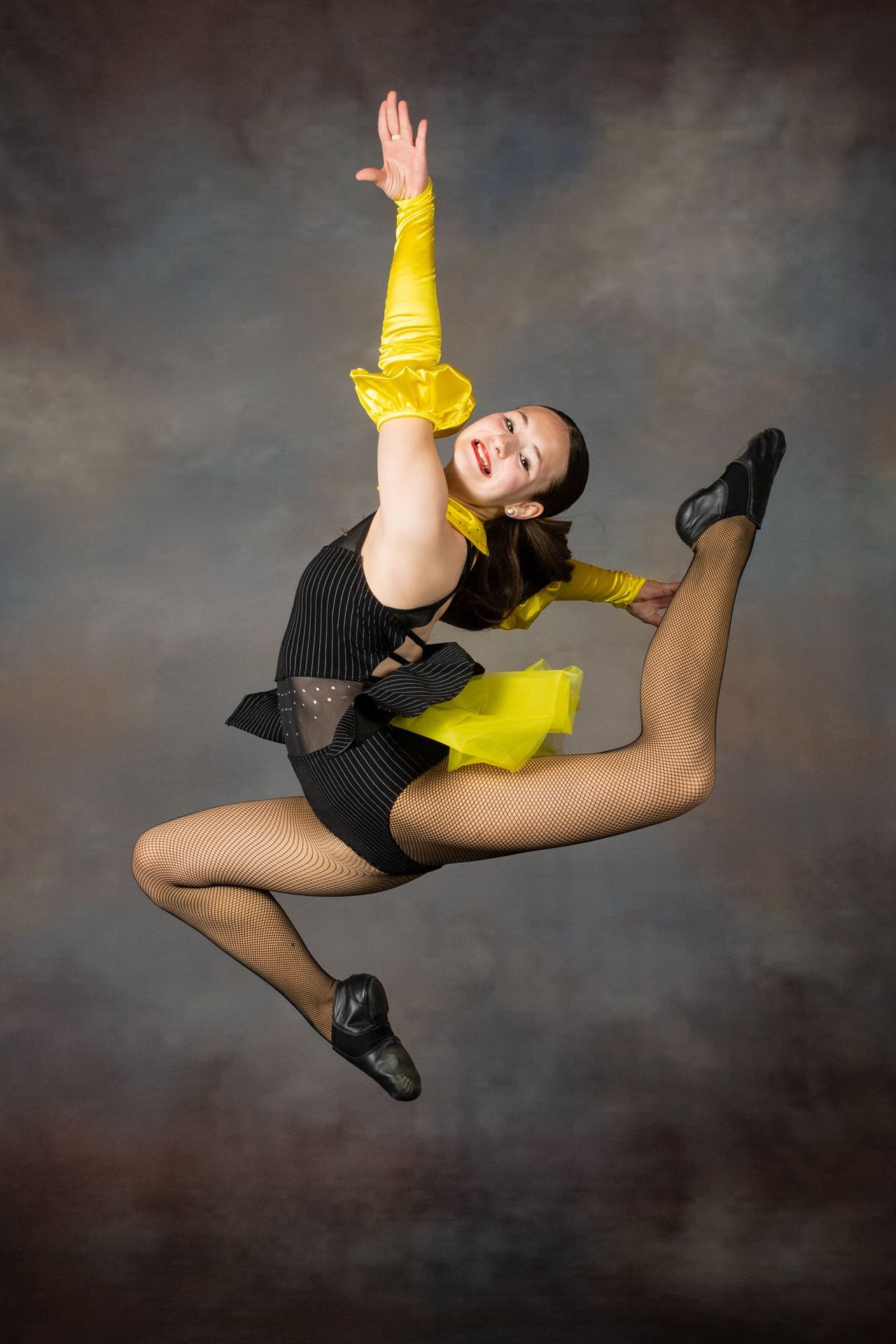 Dancer in black and yellow costume striking a high leap against a gray studio backdrop