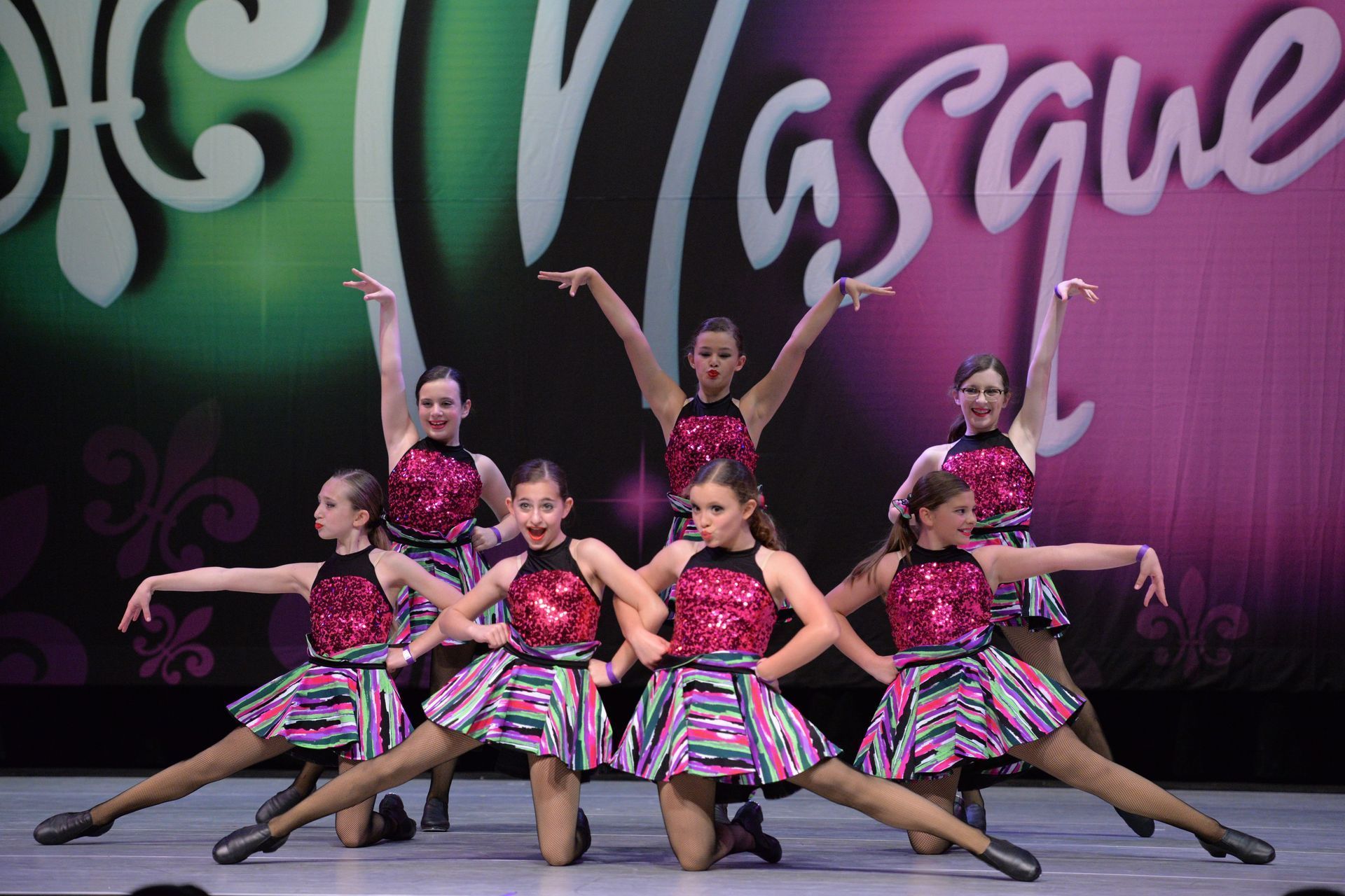 Seven dancers in pink costumes pose on stage in a synchronized routine under a purple backdrop.