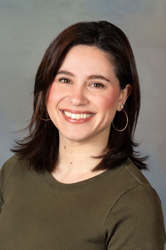 Smiling dance instructor with dark shoulder-length hair wearing an olive green top against a gray background