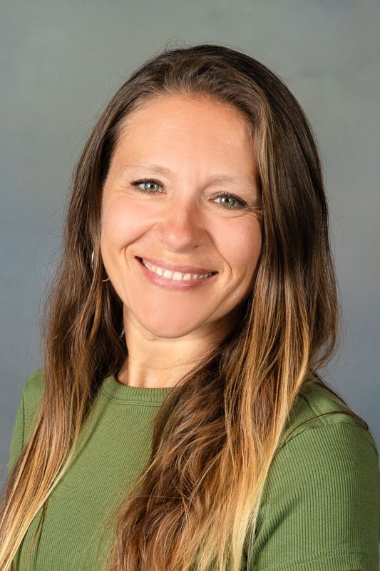 Smiling dance instructor with long brown hair in a green top against a gray background