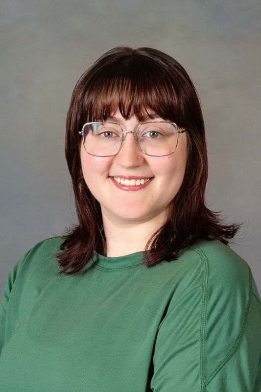 Smiling dance instructor with glasses and dark hair wearing a green shirt against a gray background
