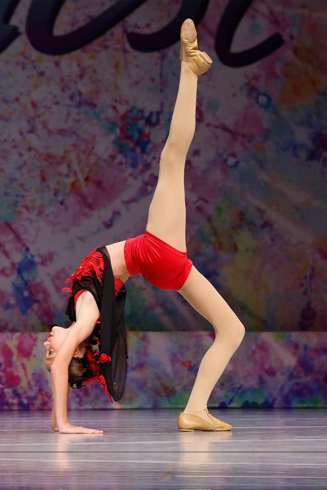 Contortionist in red shorts and black top performing a backbend with one leg raised onstage