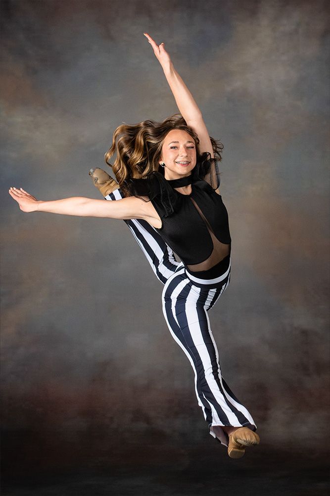 Dancer mid-leap in black-and-white striped pants against a studio backdrop, arms extended and smiling