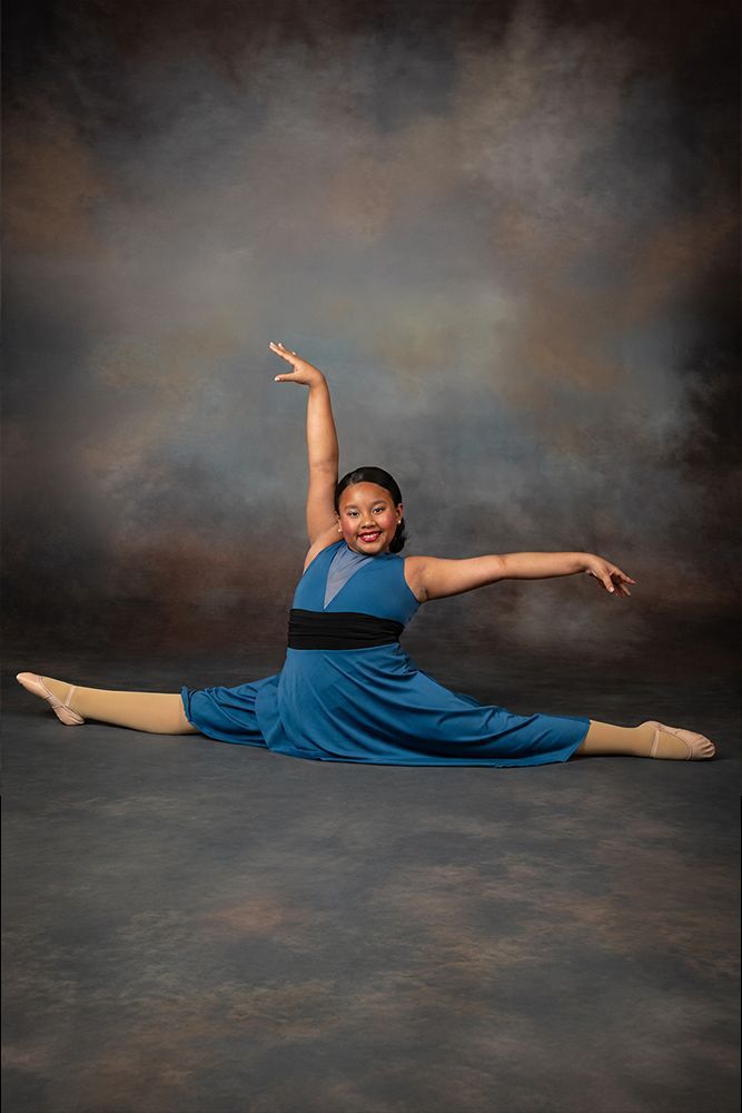 Young dancer in a blue dress doing a full split with arms raised against a studio backdrop