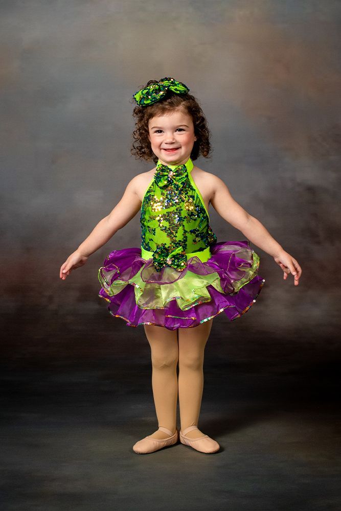 Child in a green and purple dance costume, smiling with arms outstretched against a studio backdrop.