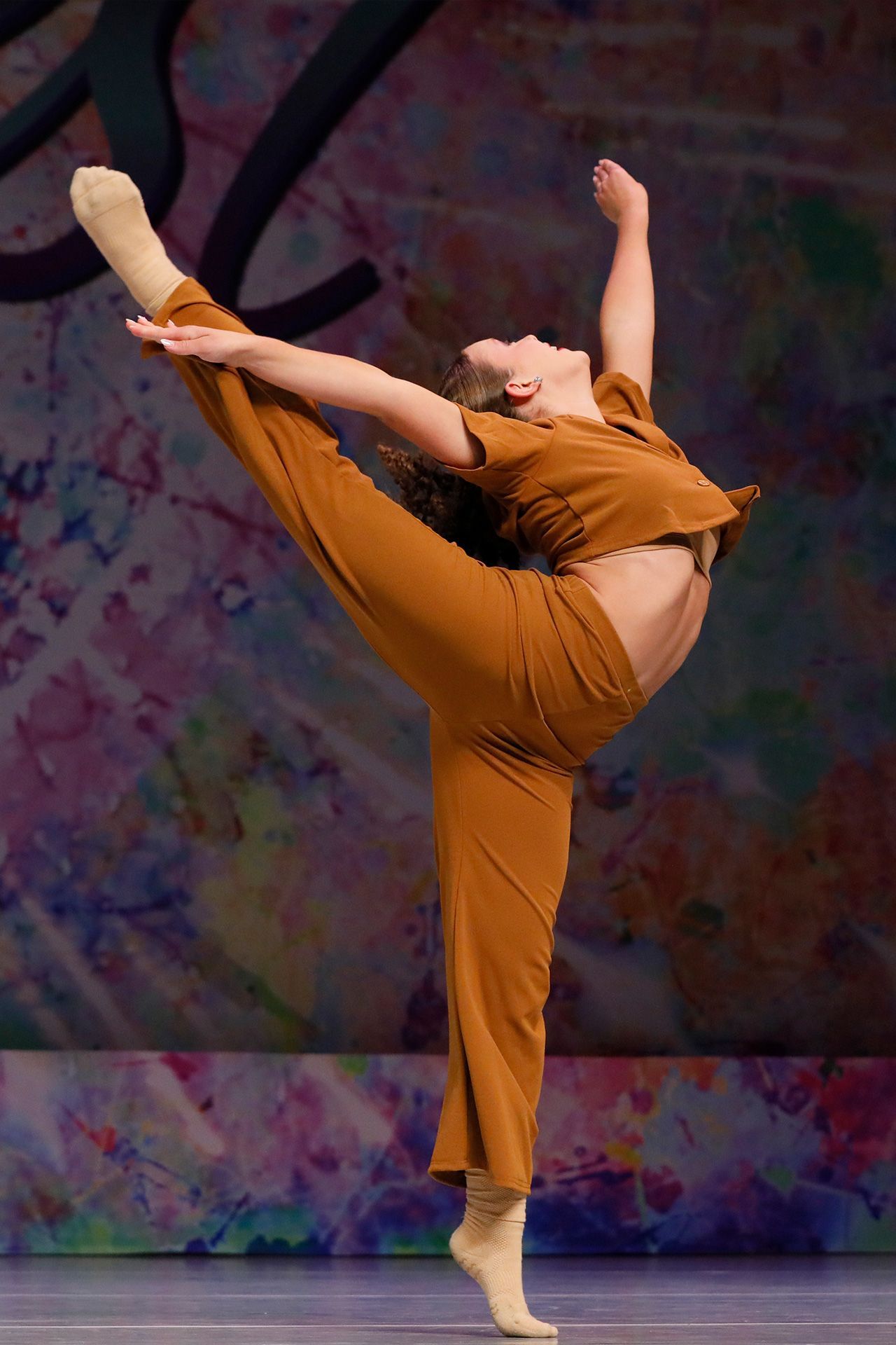 Dancer in orange performs a high leg extension onstage against a colorful backdrop