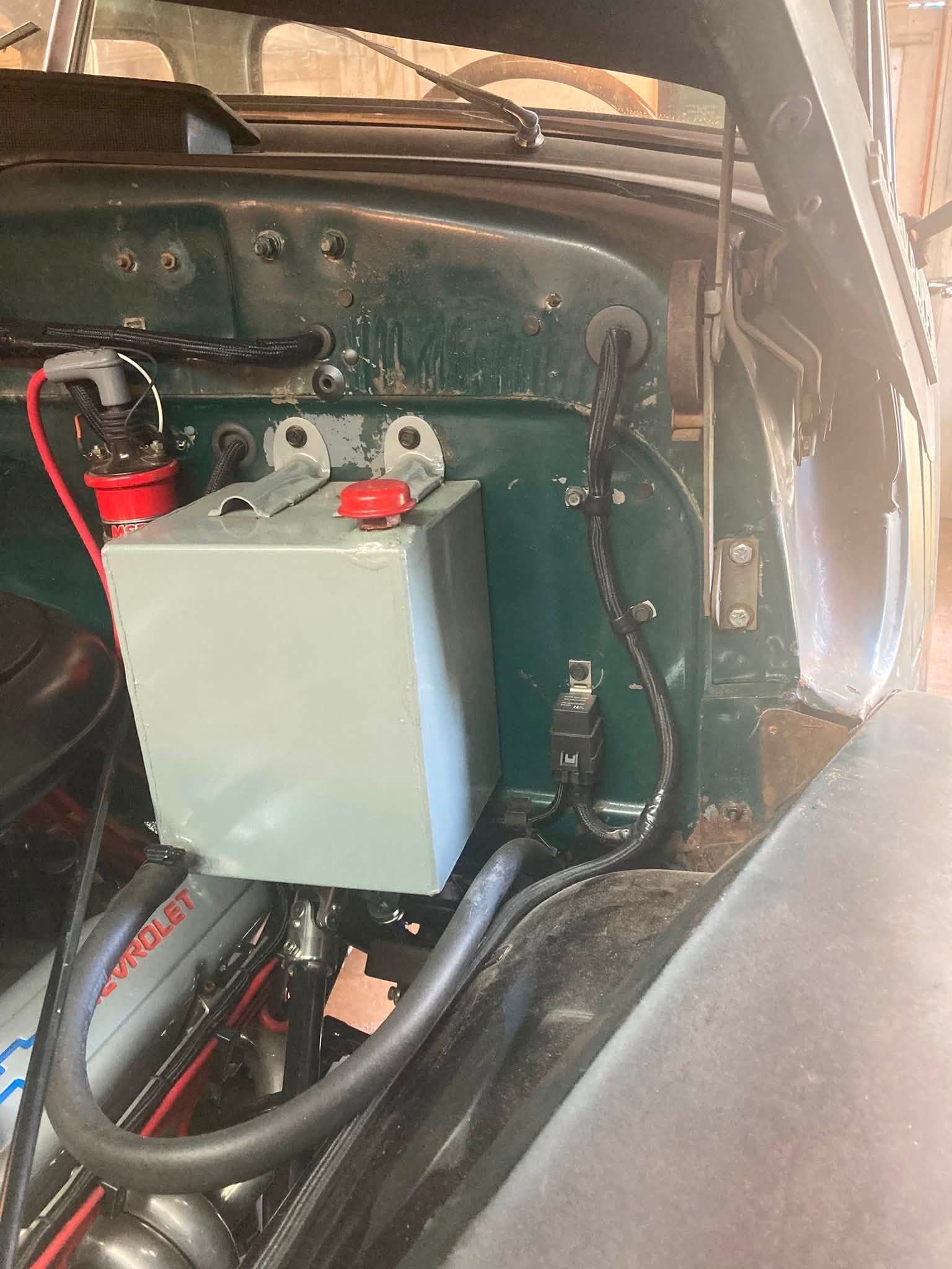 Car battery and gray power box mounted in a vehicle’s rear compartment with cables and hoses.