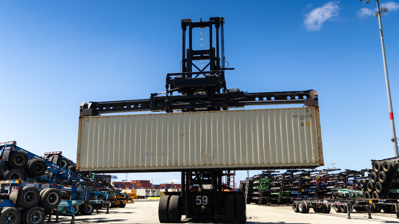 Large forklift lifting a beige shipping container in a salvage yard under a blue sky