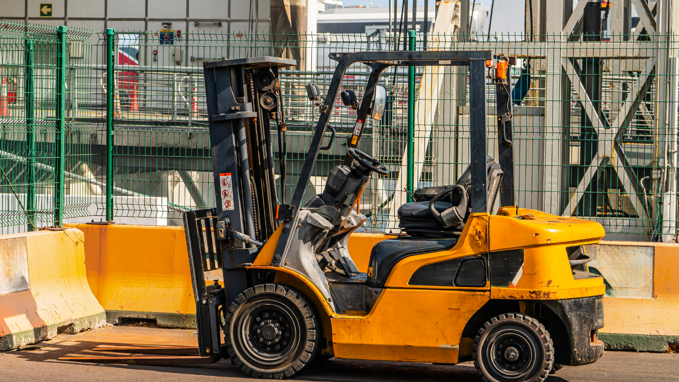 Yellow forklift parked by a concrete barrier in an industrial yard, with green fencing and equipment behind.