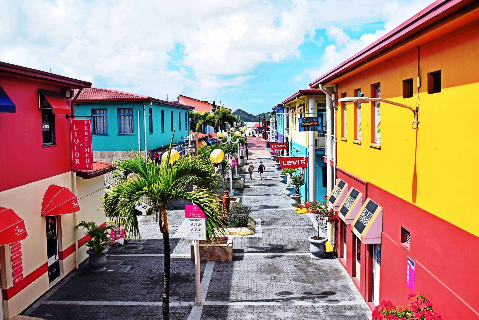 Colorful storefronts line a pedestrian street in a tropical town, with a palm tree and blue sky.