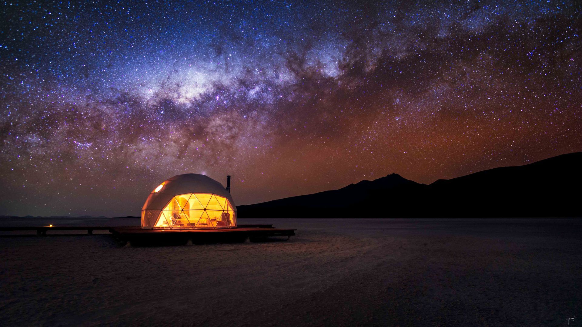 A dome tent is sitting on a dock in the middle of a lake under a starry sky.