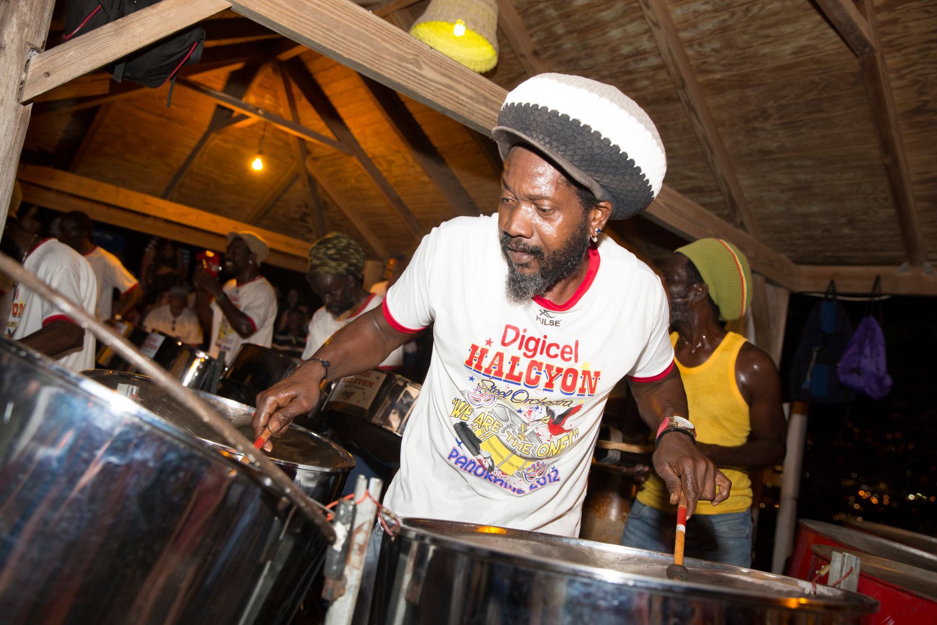 Man playing steel drums at night, wearing a white shirt and a rasta cap, with other musicians in the background.