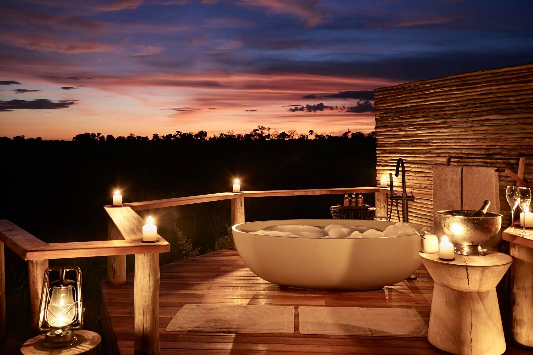 A bathtub on a wooden deck with candles and a sunset in the background