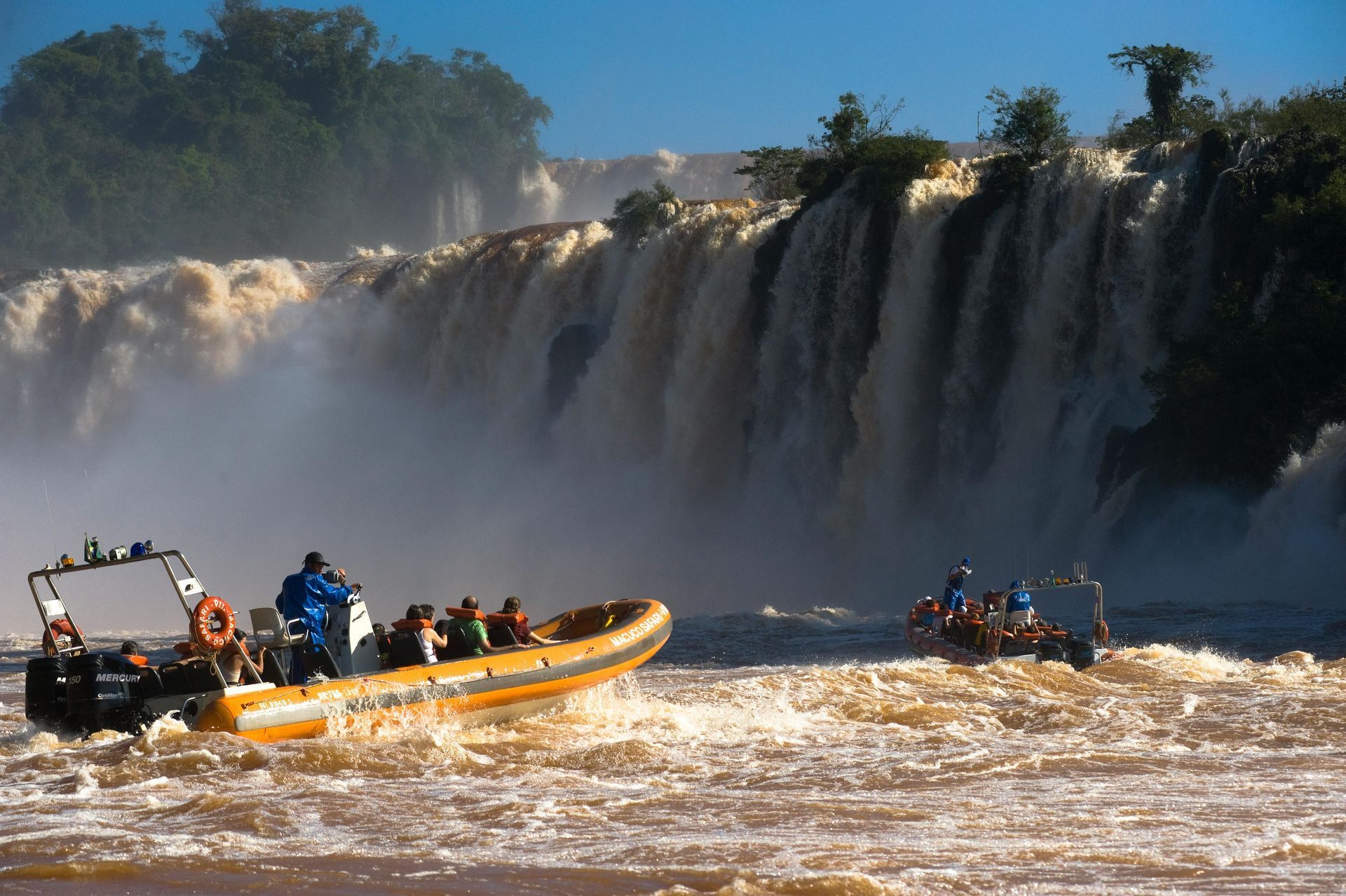 Boats near Iguazu Falls, filled with tourists, navigating turbulent water near a waterfall. Brown water and mist visible.