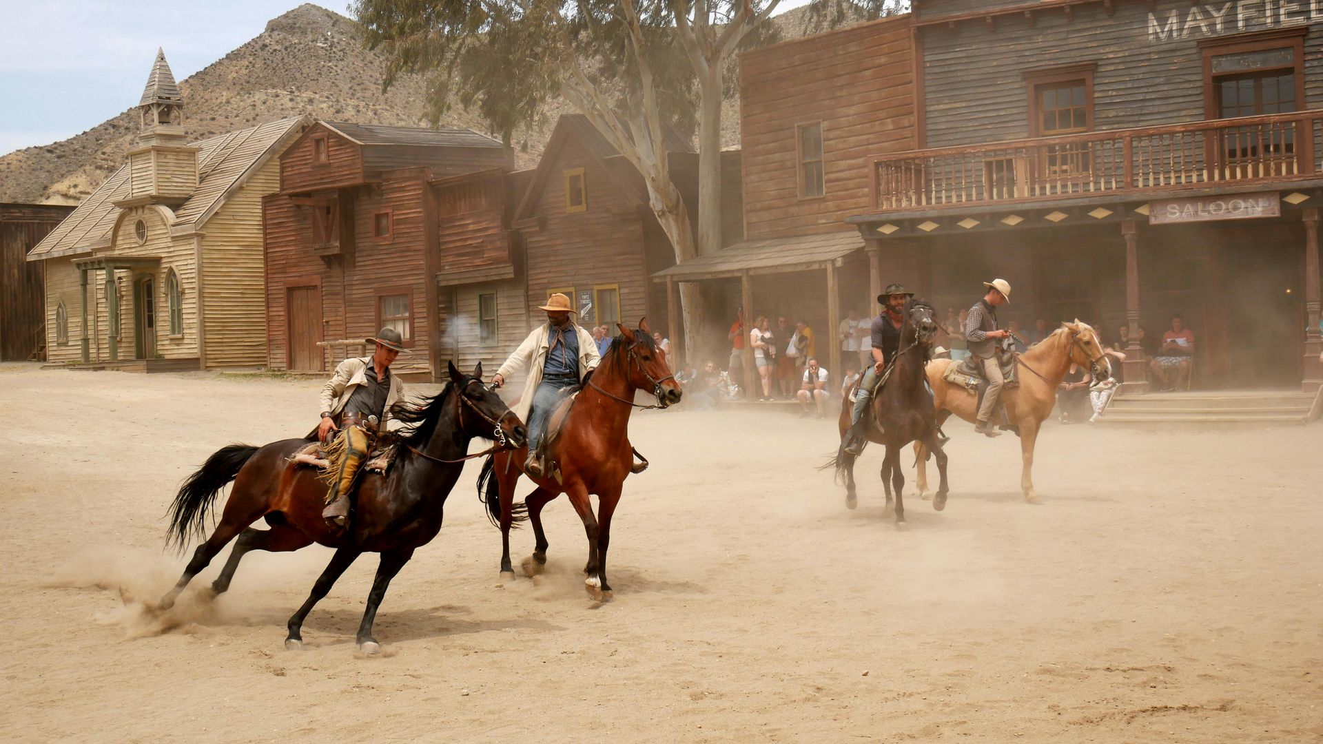 Cowboys on horseback in a dusty Wild West town, some racing, others near wooden buildings and spectators.