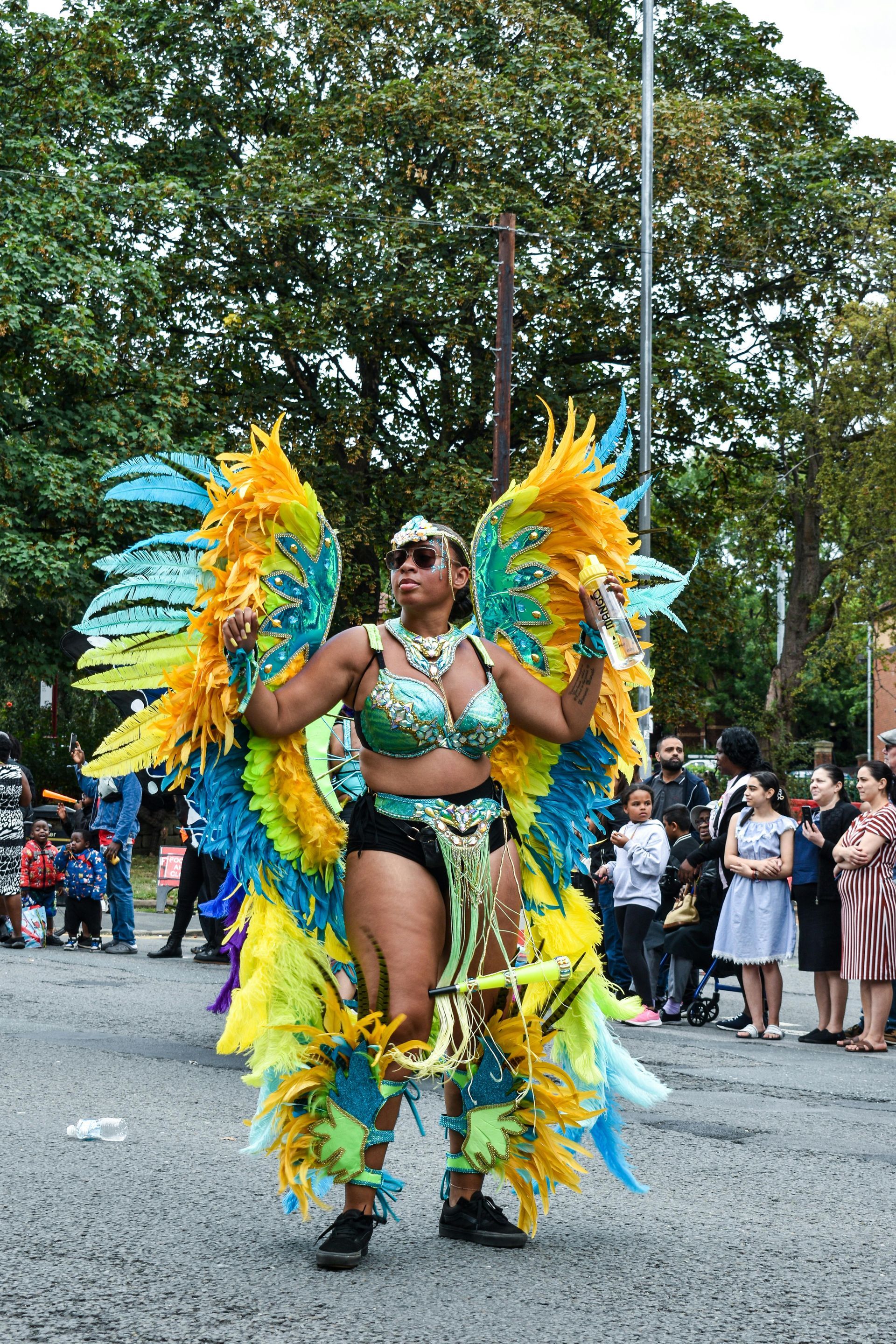 Woman in colorful carnival costume with large feather wings and headdress, dancing outdoors. People watch in background.