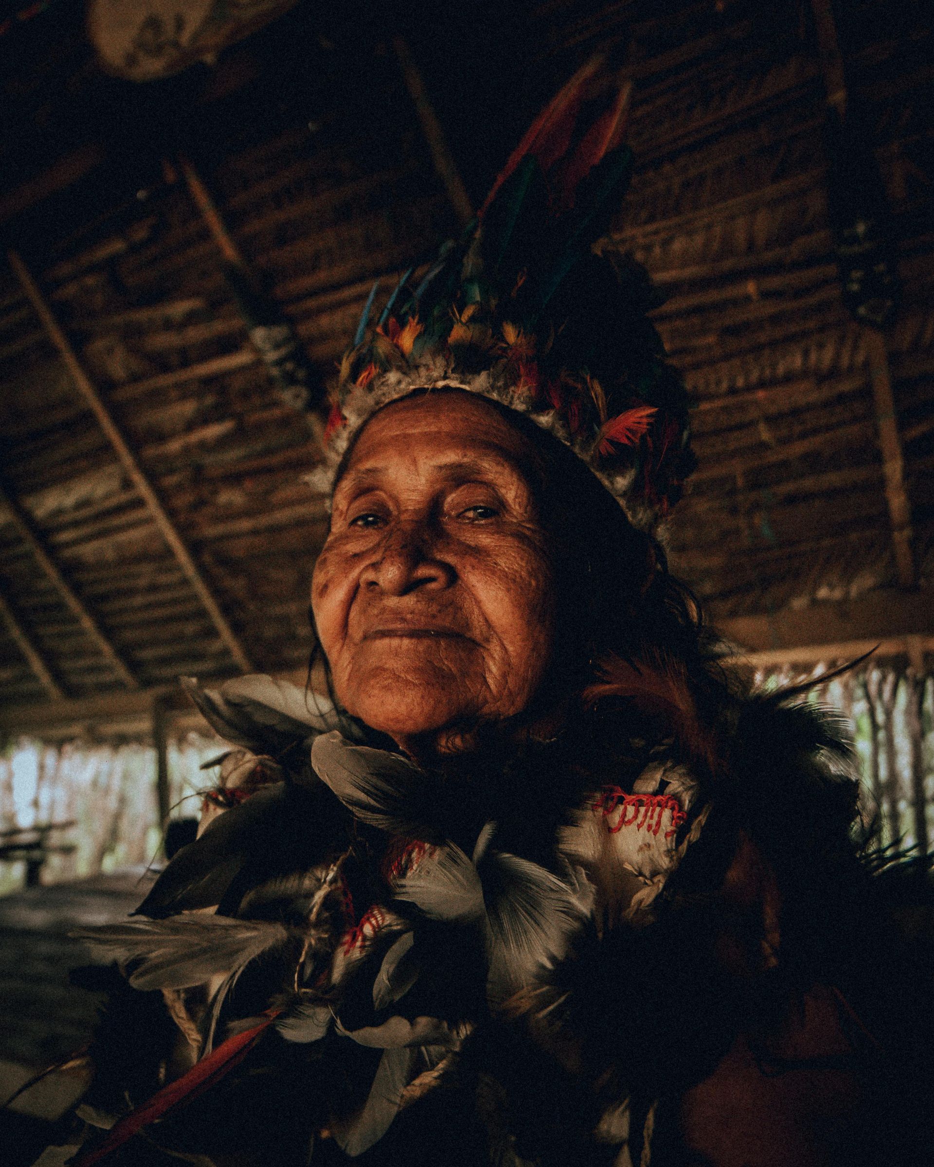An elderly Indigenous person in a feathered headdress and ceremonial attire, smiling in a dim hut.